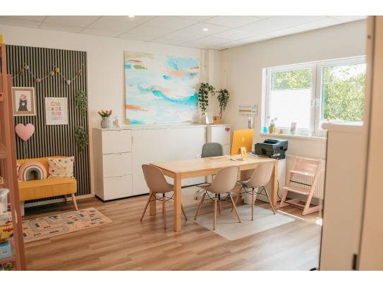 Innenraum mit Tisch, vier Stühlen, Fenster, buntem Wandbild und Sitzbereich mit Kinderbüchern