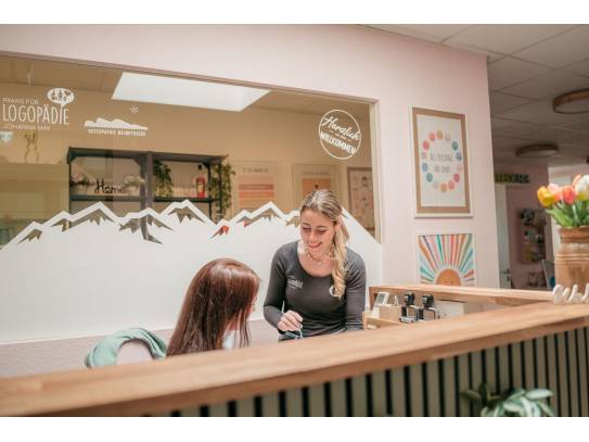 Zwei Frauen im Empfangsbereich einer Praxis, eine davon steht lächelnd hinter dem Tresen, im Hintergrund Dekoration und Beschilderung