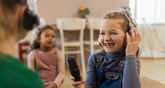 Lachendes Mädchen mit Kopfhörern hält ein Mikrofon und spricht mit einer anderen Person, im Hintergrund sitzt ein weiteres Kind – Szene in einer kindgerechten Lernumgebung