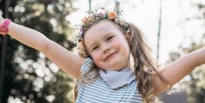 Fröhliches Mädchen mit Blumenkrone und blau-weiß gestreiftem Kleid steht mit ausgebreiteten Armen im Sonnenschein auf einer Wiese