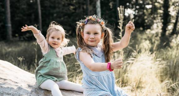 Zwei kleine Mädchen sitzen auf einem Baumstamm und strecken spielerisch die Arme in die Luft, das ältere trägt eine bunte Blumenkrone – fröhliche Szene in der Natur