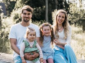 Familie May sitzt lächelnd auf einem Baumstamm in einer sonnigen Wiese – natürlicher, fröhlicher Moment im Freien