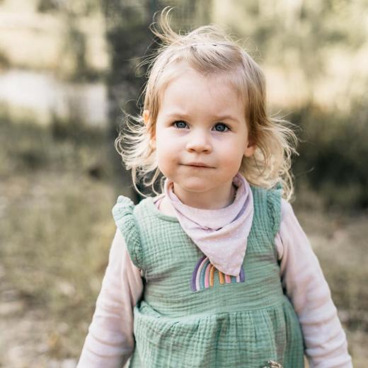 Kleines blondes Mädchen mit grünem Kleid und rosa Halstuch steht in der Natur und blickt neugierig in die Kamera