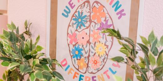 Farbiges Wandbild mit einem stilisierten Gehirn aus Blumen und dem Spruch „We all think differently“, eingerahmt von grünen Pflanzen vor rosa Wand