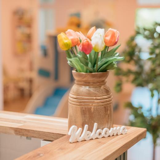 Holztheke mit Tulpenvase und Willkommensschild, dahinter ein freundlich eingerichteter Wartebereich für Kinder