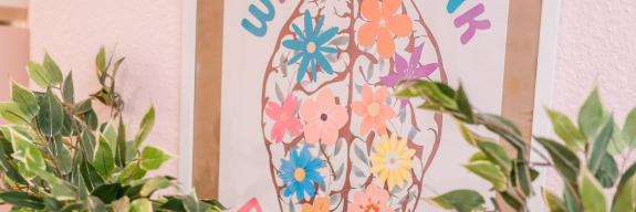 Farbiges Wandbild mit einem stilisierten Gehirn aus Blumen und dem Spruch „We all think differently“, eingerahmt von grünen Pflanzen vor rosa Wand