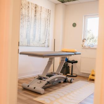 Blick in einen freundlichen Behandlungsraum mit höhenverstellbarer Therapieliege, Naturmotiv an der Wand, Deckenbild mit Baumkrone und Tageslichtfenster