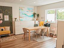 Innenraum mit Tisch, vier Stühlen, Fenster, buntem Wandbild und Sitzbereich mit Kinderbüchern