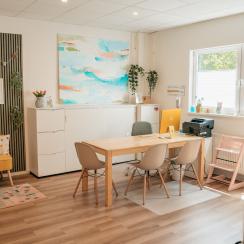 Innenraum mit Tisch, vier Stühlen, Fenster, buntem Wandbild und Sitzbereich mit Kinderbüchern