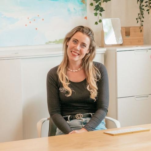 Freundliche Frau mit langen blonden Haaren sitzt lächelnd hinter einem hellen Holzschreibtisch in einer modern eingerichteten Praxis für Logopädie